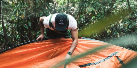 Eine Person, die eine Mütze trägt, baut ein orangefarbenes Zelt in einem üppigen grünen Wald auf, umgeben von Blättern und Bäumen.