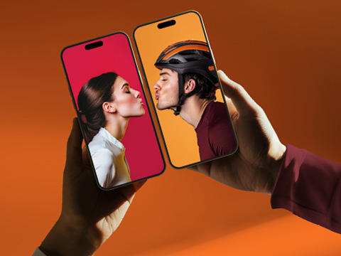 Zwei Hände halten Smartphones, auf deren Bildschirmen eine Frau und ein Mann im Profil zu sehen sind, die sich vor rotem und orangefarbenem Hintergrund scheinbar über die Bildschirme hinweg küssen.