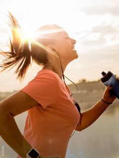 Eine Frau in einem rosa Shirt, die bei Sonnenaufgang im Freien joggt, lächelt, Kopfhörer trägt und eine Wasserflasche in der Hand hält, während hinter ihr die Sonne scheint.