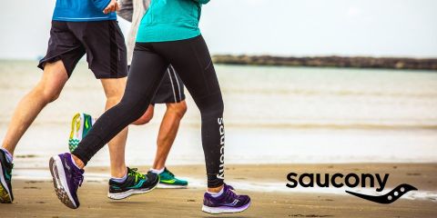Drei Personen laufen in Sportkleidung an einem Sandstrand entlang; in der rechten unteren Ecke ist das Saucony-Logo zu sehen.
