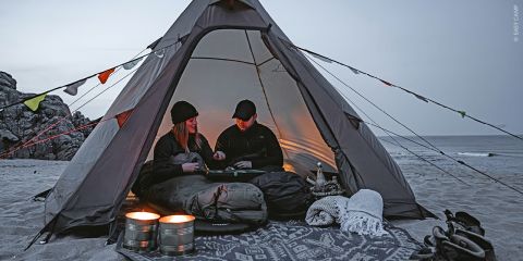 Zwei Menschen sitzen in der Abenddämmerung in einem Zelt am Strand und spielen ein Brettspiel; daneben liegen Kerzen, Decken und Campingausrüstung.