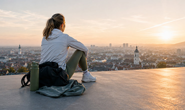 Eine Person sitzt bei Sonnenaufgang auf einem Dach und blickt auf eine Stadtlandschaft mit Gebäuden und fernen Hügeln; neben ihr liegen eine Wasserflasche und eine Jacke.