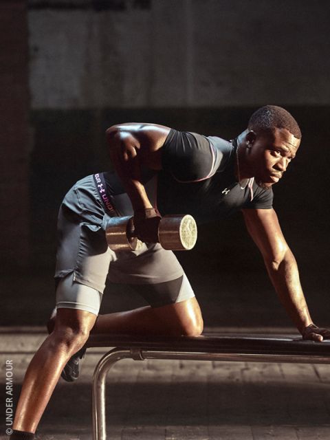 Ein Mann in Sportkleidung stemmt eine Hantel, während er sich auf eine Bank stützt; konzentriert trainiert er in einem schwach beleuchteten Fitnessstudio.