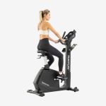 Frau in Sportkleidung auf einem stationären Heimtrainer, Blick nach rechts, vor einem schlichten weißen Hintergrund.