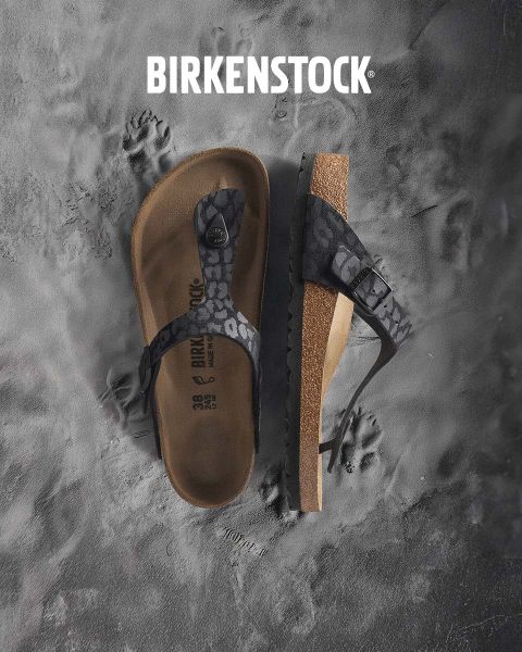 Eine Person in beigen Birkenstock-Sandalen steht neben einem Wasserspiel in einer üppigen Gartenlandschaft. Der Bereich ist von Grün umgeben und das Wort „BIRKENSTOCK“ ist deutlich über dem Bild zu sehen.