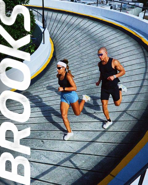 Eine Gruppe von fünf Personen rennt energisch über einen Zebrastreifen. Sie tragen farbenfrohe Sportkleidung, darunter Shorts und Tanktops. Die Szene ist lebhaft, über ihnen prangt deutlich das Wort „BROOKS“.