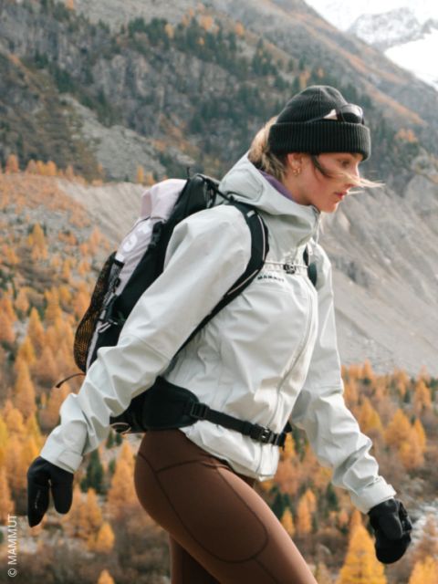 Eine Frau wandert im Herbst in der freien Natur, bekleidet mit einer Jacke, einer Mütze, Handschuhen und einem Rucksack. Im Hintergrund sind Berge und Orangenbäume zu sehen.