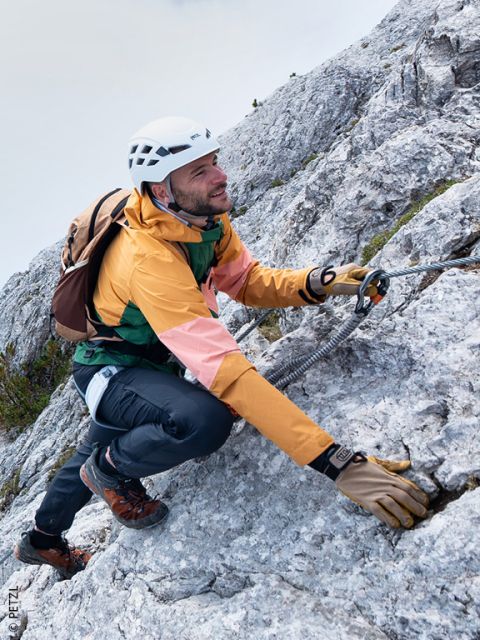 Ein Mann in Kletterausrüstung steigt einen felsigen Berghang hinauf und hält sich dabei mit einem Karabiner an einem Sicherheitsseil fest.