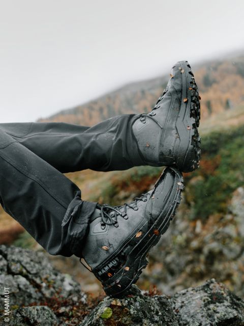 Eine Person in schwarzen Wanderstiefeln, an denen Schmutz und Blätter haften, ruht ihre Füße im Freien auf Felsen aus, im Hintergrund ist eine verschwommene Berglandschaft zu sehen.
