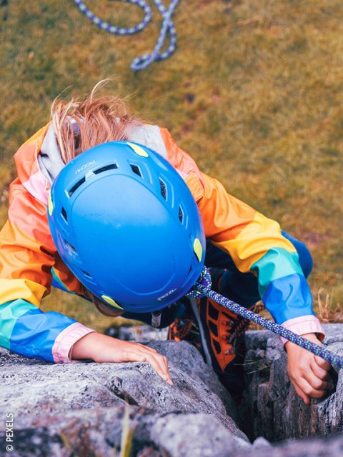 Eine Person mit blauem Helm und bunter Jacke klettert an einer Felswand empor, gesichert durch ein Seil. Blick von oben, darunter ist ein grasbewachsener Boden zu sehen.