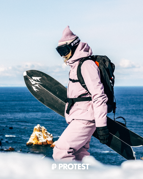 Eine Person in einem rosa Schneeanzug und einer Mütze, die ein Snowboard in der Hand hält, steht am Meer, im Hintergrund sind Felsformationen und unten das Wort „PROTEST“ zu sehen.