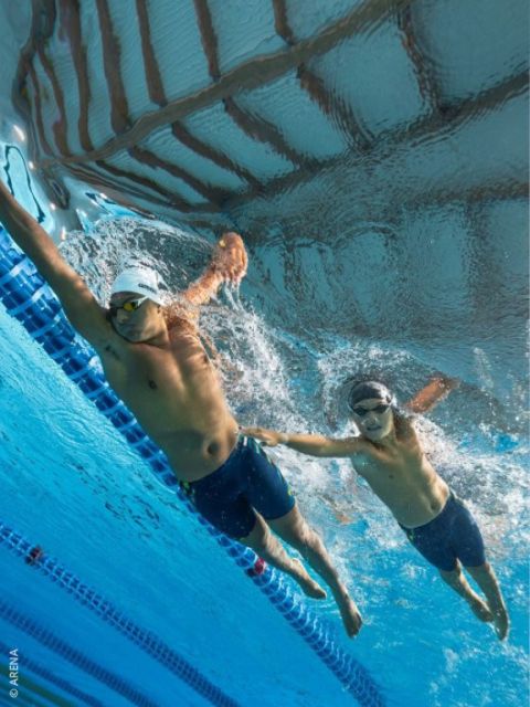 Zwei Schwimmer mit Schwimmbrillen und Badekappen liefern sich in einem Schwimmbecken ein Unterwasserrennen; einer streckt in der Nähe der Bahntrennung die Hand nach dem anderen aus.