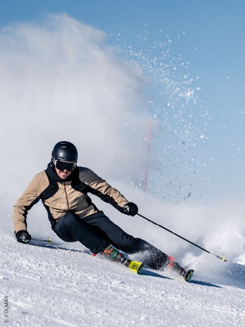 Ein Skifahrer mit Helm und hellbrauner Jacke zieht scharfe Schwünge auf einem verschneiten Hang und wirbelt dabei unter strahlend blauem Himmel Pulverschnee auf.