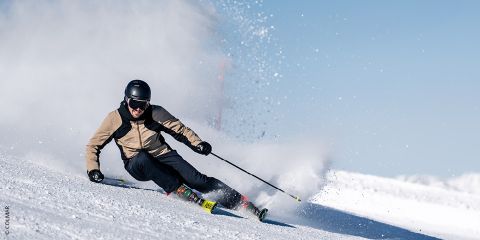 Ein Skifahrer in einer beigen Jacke und einem schwarzen Helm zieht scharfe Schwünge auf einem verschneiten Hang und wirbelt dabei eine Schneewolke gegen den strahlend blauen Himmel auf.