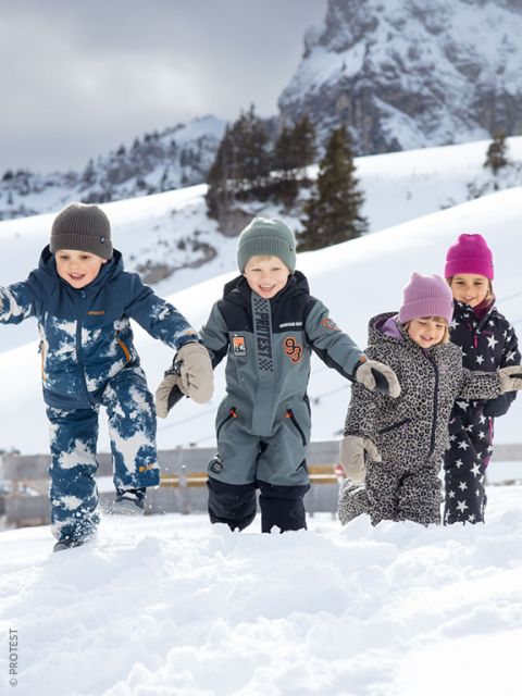 Vier Kinder in Winterkleidung spielen und lächeln im Schnee, im Hintergrund sind schneebedeckte Berge und Bäume zu sehen.