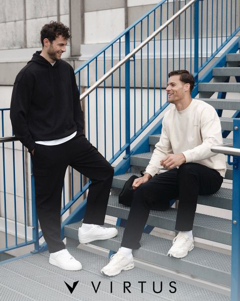 Zwei Männer in Sportkleidung von Virtus, der eine in einem hellgrauen Kapuzenpulli mit schwarzem Seesack, der andere in einer schwarzen Jacke, lächeln und gehen zusammen in einer modernen Halle spazieren. Das Virtus-Logo ist über ihnen zu sehen.
