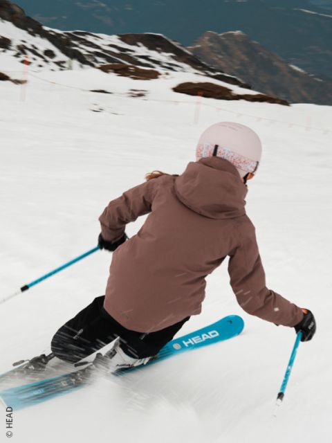 Ein Skifahrer in beiger Jacke, blauer Hose und weißem Helm fährt auf einer verschneiten Piste bergab, macht eine scharfe Kurve und wirbelt Schnee auf. Im Hintergrund sind Berge zu sehen.