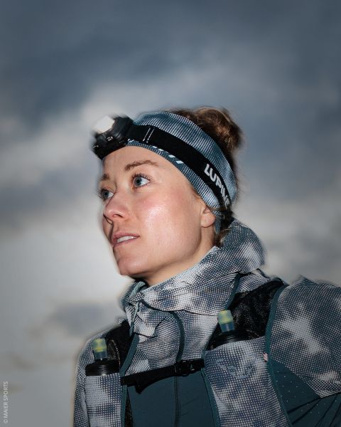 Eine Frau, die eine Stirnlampe, ein gemustertes Stirnband und eine Trailrunning-Weste trägt, blickt gegen den bewölkten Himmel empor.