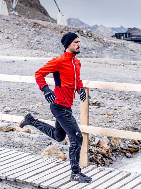 Ein Mann in einer roten Jacke, einer schwarzen Hose, Handschuhen und einer Mütze läuft auf einer Holzbrücke in einer felsigen, bergigen Gegend.