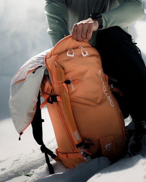 Ein Wanderer öffnet einen orangefarbenen Ortovox-Rucksack in einer verschneiten Berglandschaft.