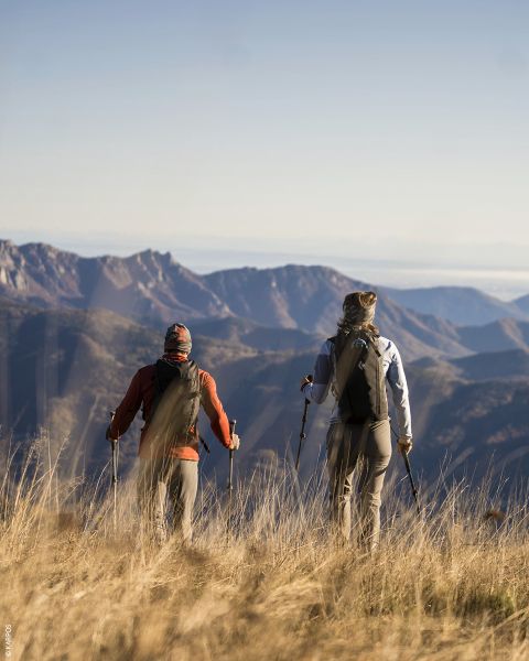 Zwei Wanderer mit Rucksäcken und Wanderstöcken stehen im hohen Gras und blicken unter klarem Himmel auf ferne Bergketten.